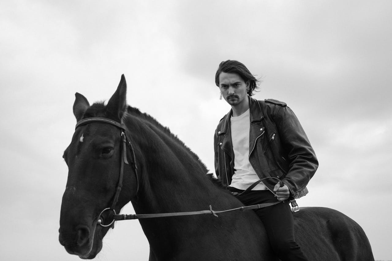 A stylish monochrome image of a man riding a black horse outdoors, wearing a leather jacket.
